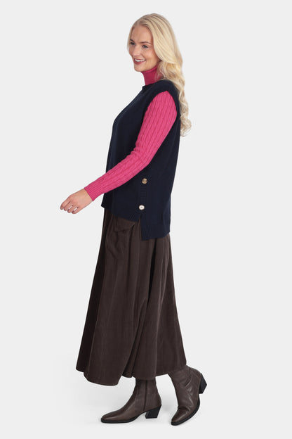 Woman wearing a navy vest over a pink sweater with a brown skirt and boots on a white background
