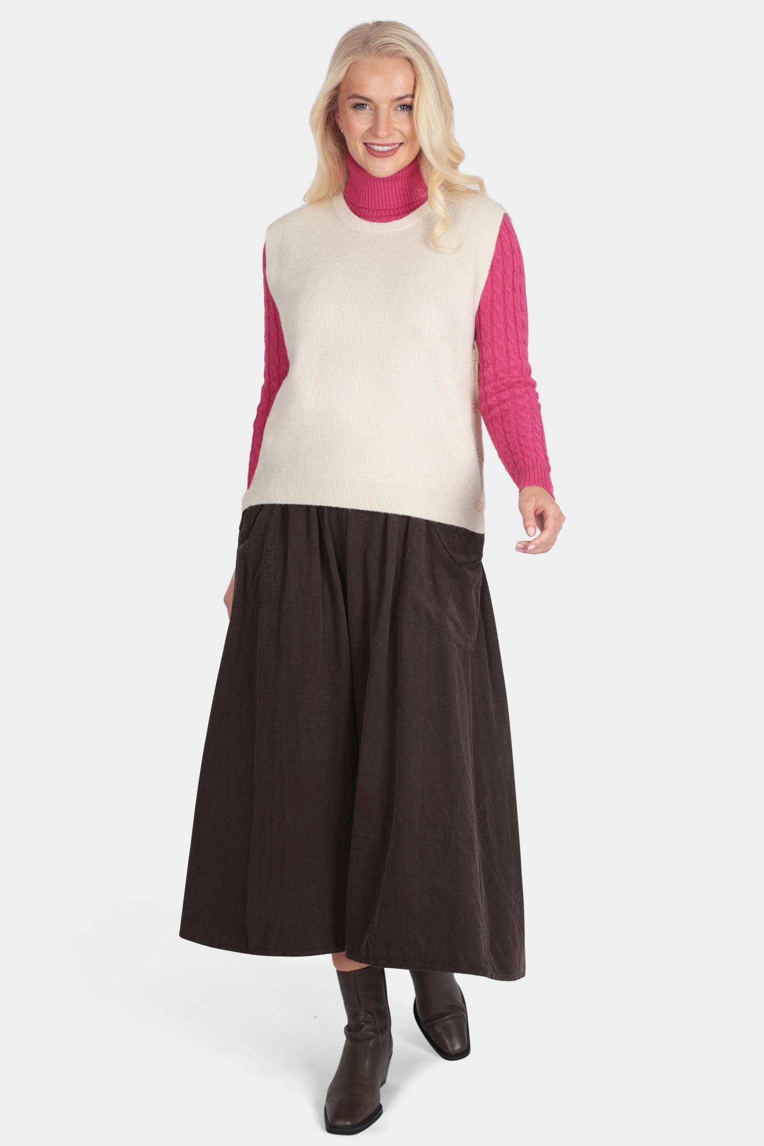 Woman wearing a cream vest over a pink shirt with a brown skirt on a white background