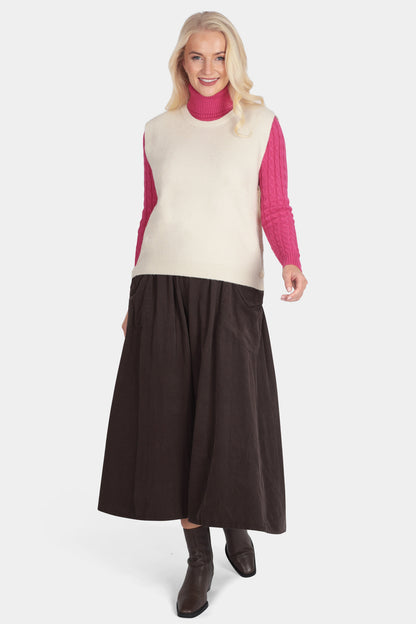 Woman wearing a cream vest over a pink shirt with a brown skirt on a white background