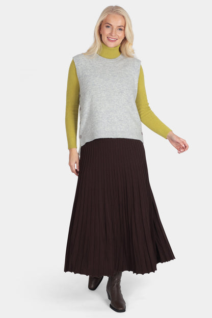 Woman wearing a sleeveless gray top, green long-sleeve shirt, and brown pleated skirt on a white background