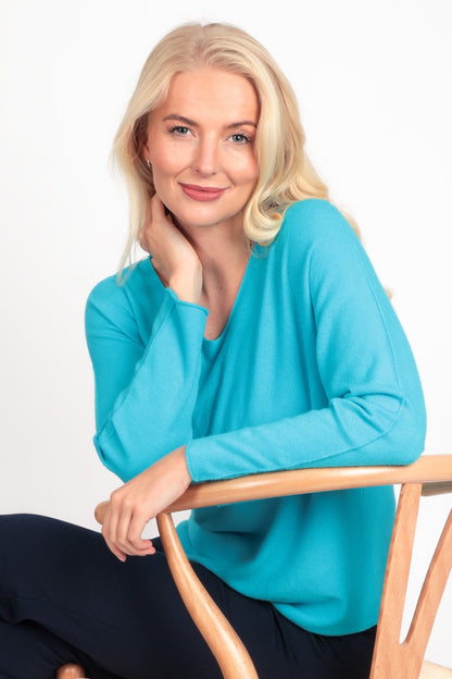 Woman sitting on a wooden chair wearing a turquoise sweater and black pants against a white background