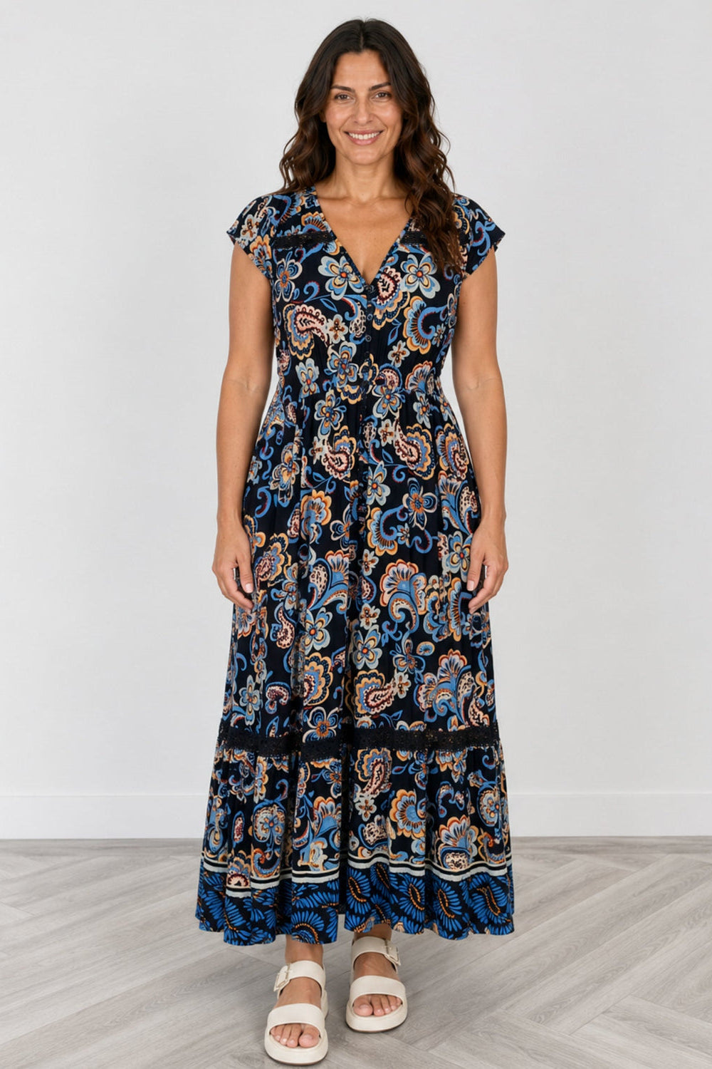 A woman modeling a navy blue maxi dress with a detailed paisley pattern in blue, orange, and beige, paired with white slide sandals, set against a neutral studio background.