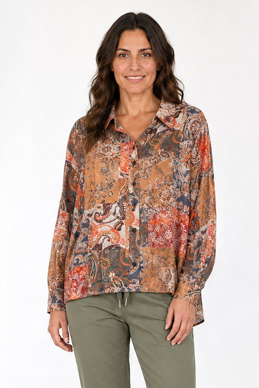 A woman modeling a long-sleeve button-up shirt with a dense abstract pattern in orange, brown, and beige, paired with olive green cropped pants and white sneakers, set against a clean white studio background.